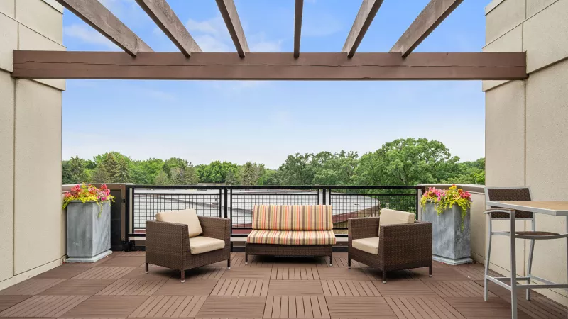 Outdoor patio with a brown wicker sofa, two armchairs, and flowering planters under a wooden pergola, overlooking a green landscape.