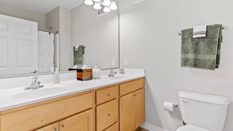 Modern bathroom with a light wood double vanity, two sinks, large mirror, toilet, and green towels on a rack.