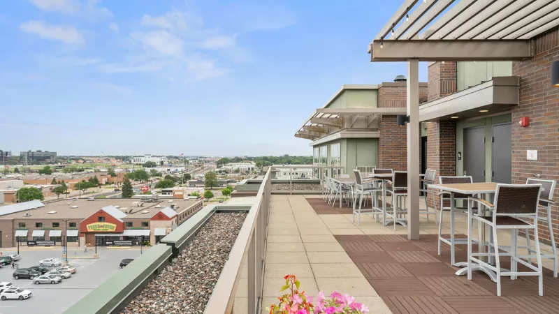 Rooftop patio with high-top tables, chairs, and potted flowers overlooking a city skyline under a blue sky.