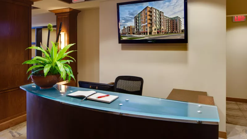 Reception desk with curved wood and blue glass top, plant, book, and a wall-mounted TV displaying a modern building.