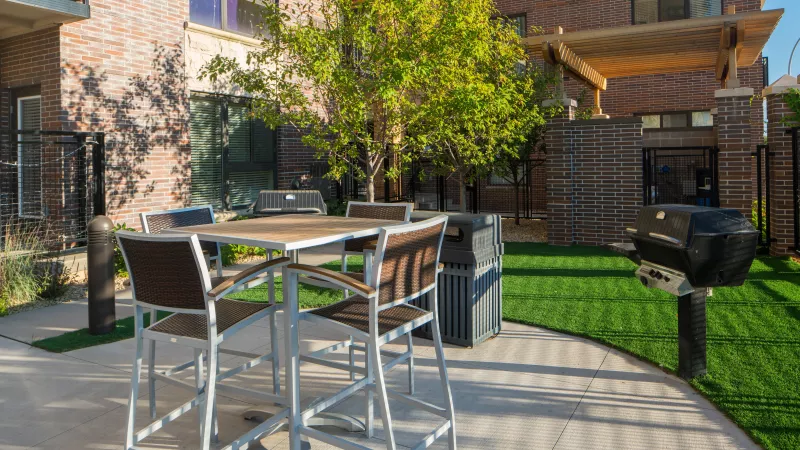 Sunny outdoor patio with a high-top table, chairs, barbecue grill, and artificial turf, backed by a brick building.
