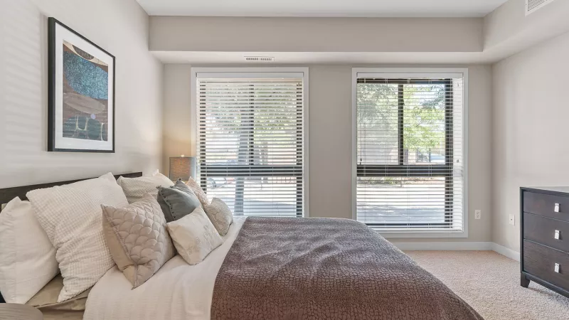 Bright bedroom with a bed, white and beige pillows, a brown blanket, and two large windows with blinds. Dark dresser.