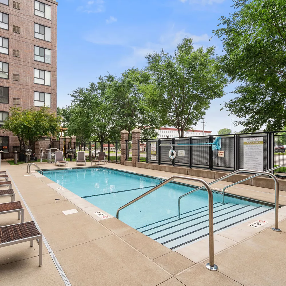 Outdoor swimming pool with lounge chairs next to a modern brick apartment building and green trees under a blue sky.