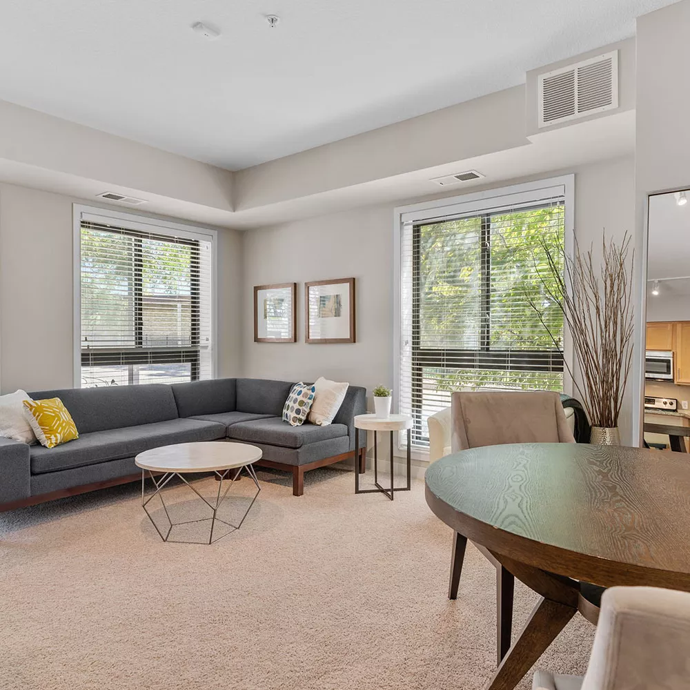 Bright living room with gray sectional, round coffee table, and dining area. Large windows and light colored walls.