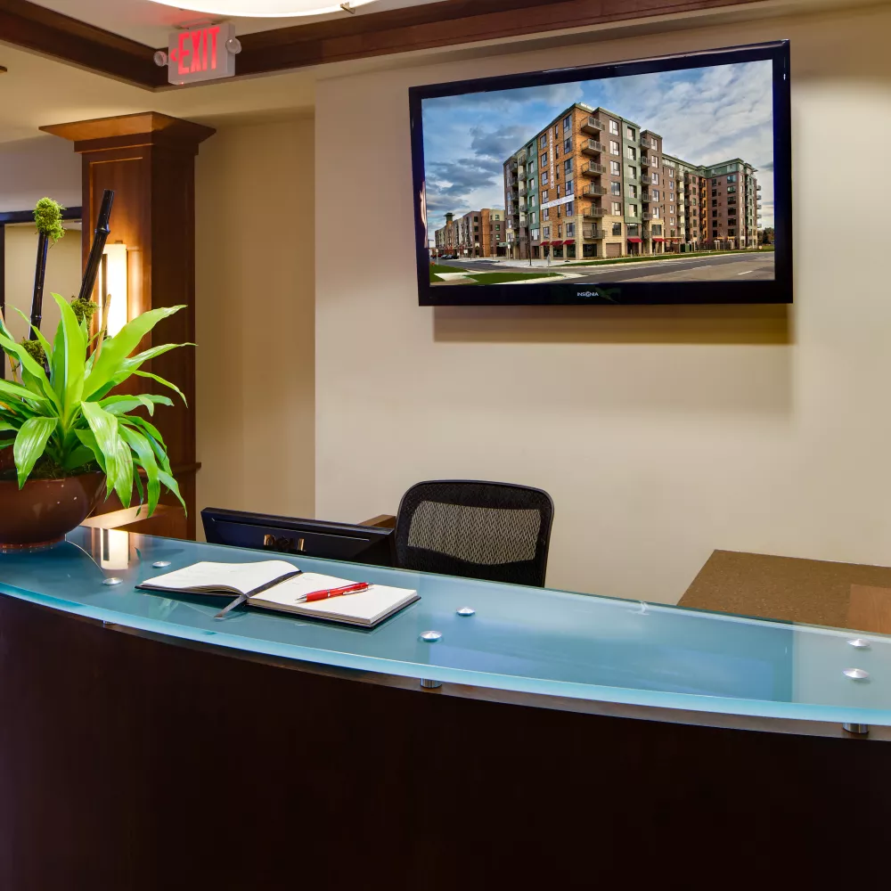Reception desk with curved wood and blue glass top, plant, book, and a wall-mounted TV displaying a modern building.