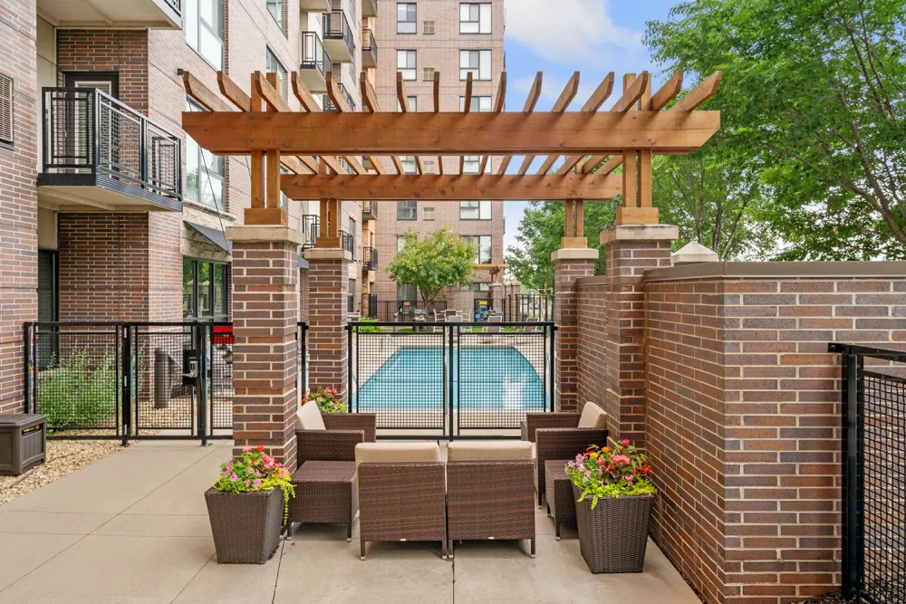 Outdoor patio with brown wicker furniture, wooden pergola, and potted flowers. A swimming pool and apartment buildings in background.